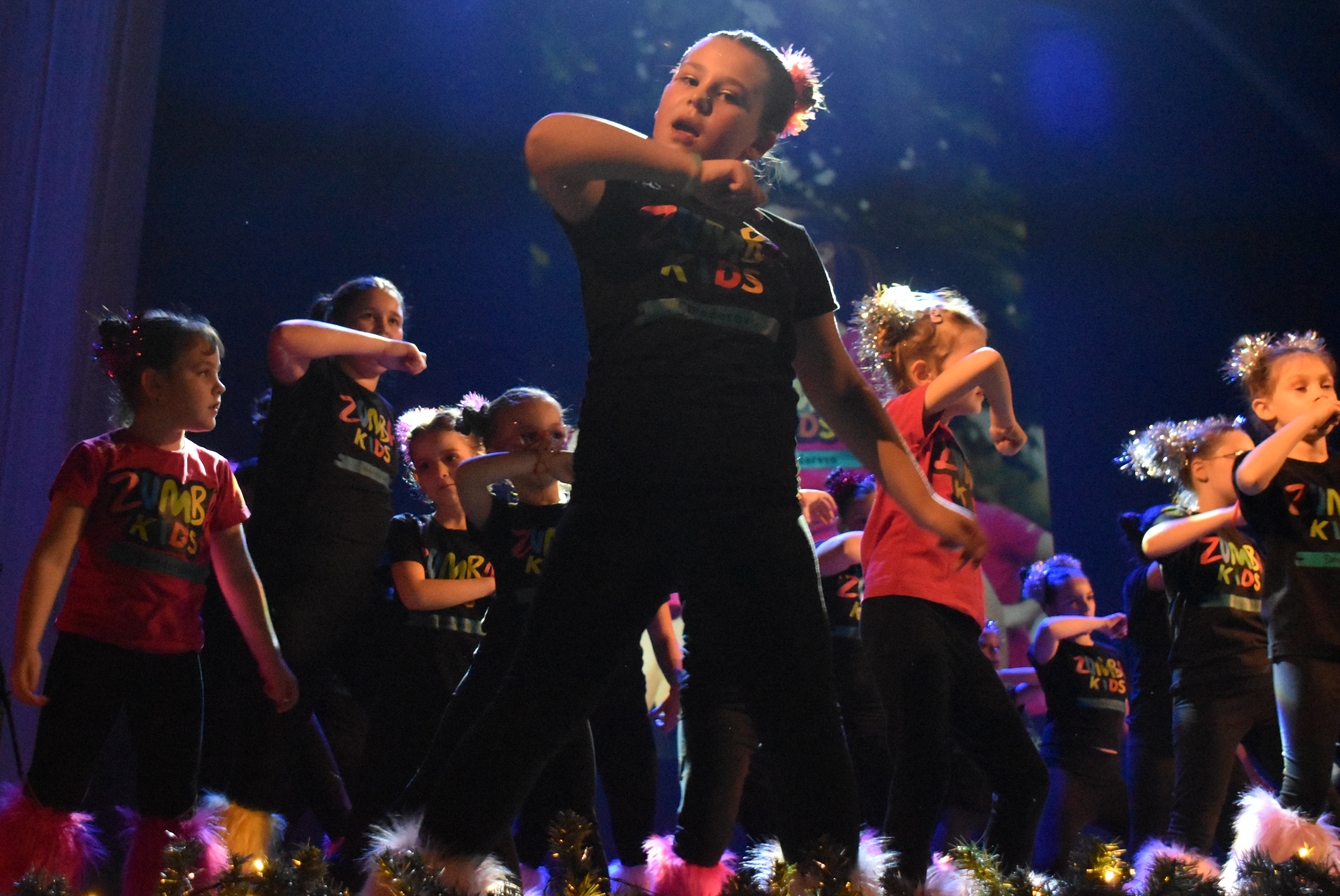 Deca u Zumba Kids majicama plešu na sceni pod plavim svetlom / Children in Zumba Kids shirts dancing on stage under blue light